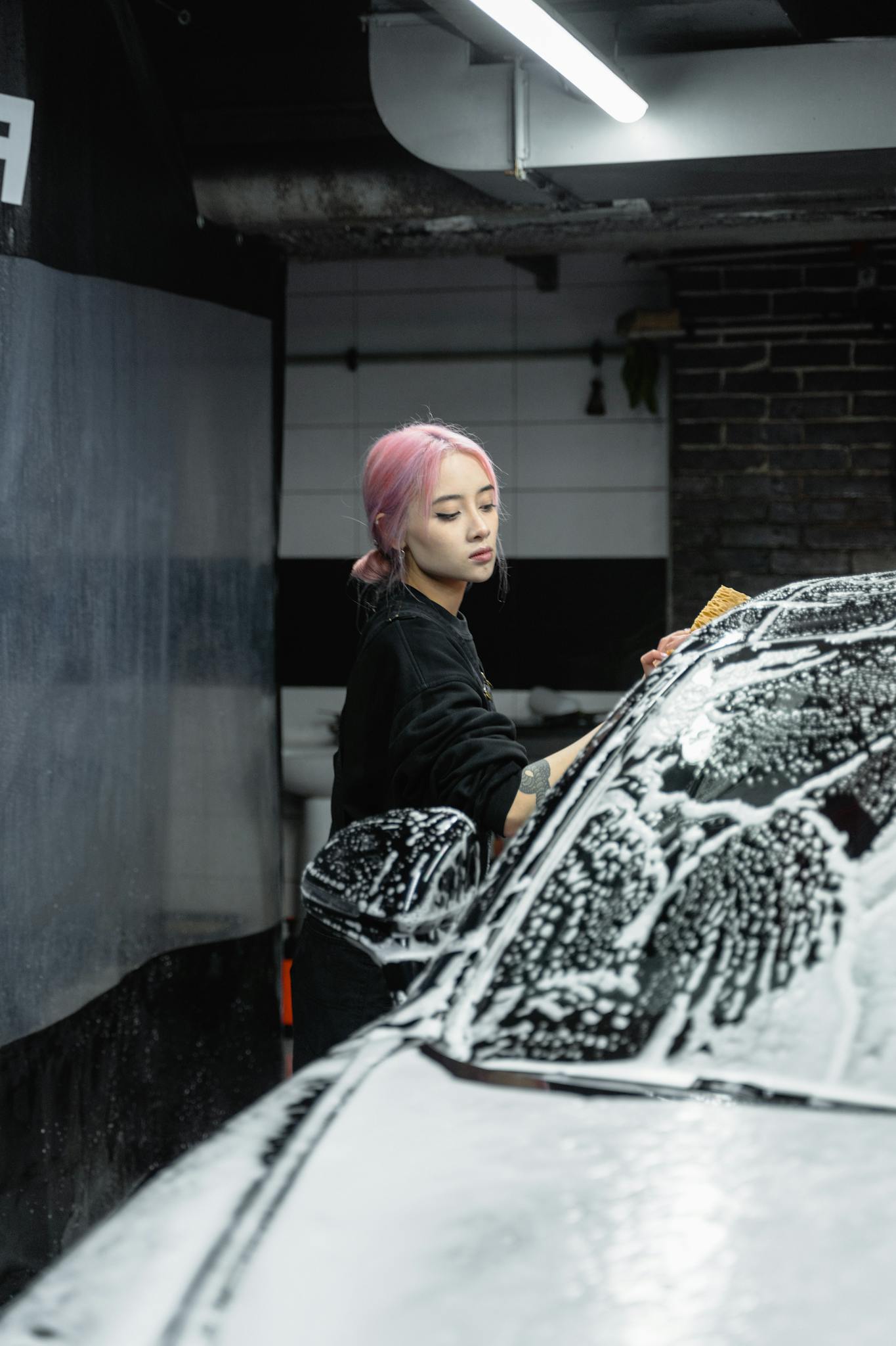 Woman with pink hair washing a car with soap in a garage setting.